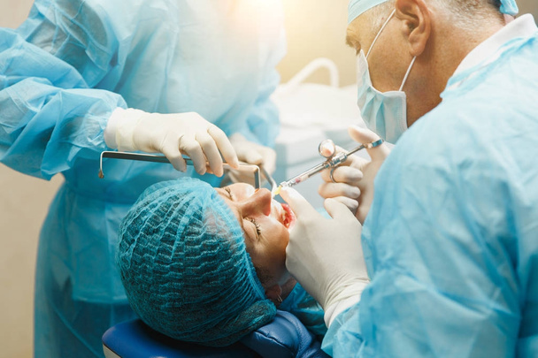 Hombre viejo cirujano dentista profesional en uniforme y asistente femenino ayuda a realizar la operación instalar dientes de implante dental de la mujer paciente en la oficina de luz clínica con equipo de herramientas modernas
. - Foto, Imagen
