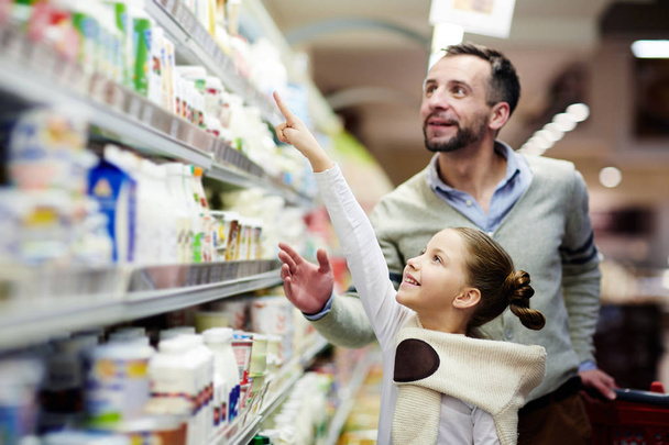 Happy girl pointing at pack of youghurt on upper shelf in supermarket - Фото, изображение