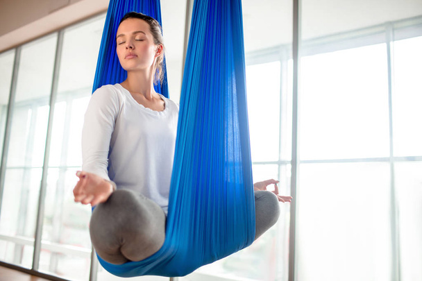 Full length portrait of pretty young woman with closed eyes sitting in lotus position and meditating while practicing anti-gravity yoga at spacious fitness club - Photo, Image