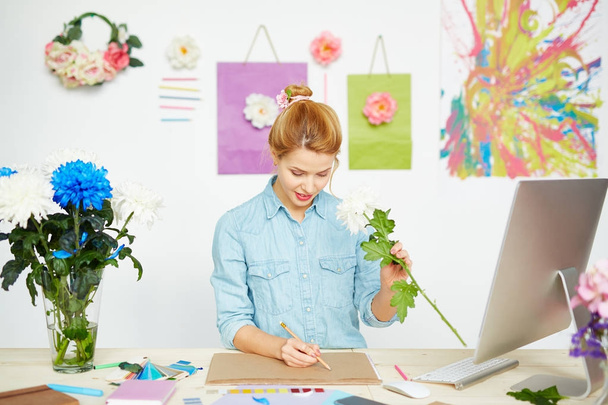 Mooie kunstenaar met rommelig haar broodje maken schets van witte chrysanten met belangstelling terwijl zittend in mooie studio met bloemen ingericht - Foto, afbeelding