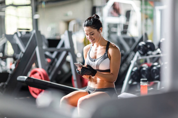 Portrait of smiling slim woman listening to music using smartphone while sitting on bench among machines before work out  in gym - Photo, Image