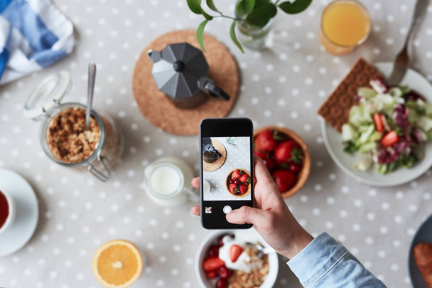 Foodbloggerin, die mit Hilfe des Mobiltelefons das appetitliche Frühstück fotografiert, konzentriert sich auf den Vordergrund - Foto, Bild