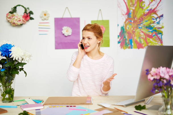 Annoyed female owner of design studio talking to her business partner on smartphone and gesticulating at the same time, abstract painting and flower decorations hanging on wall - Photo, image