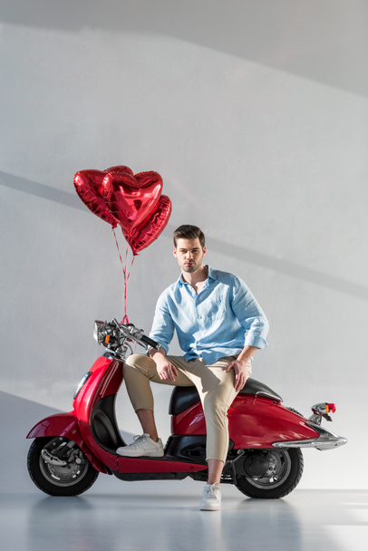 νεαρός άνδρας με μπαλόνια σε σχήμα καρδιάς που κάθεται στο κόκκινο σκούτερ - Φωτογραφία, εικόνα