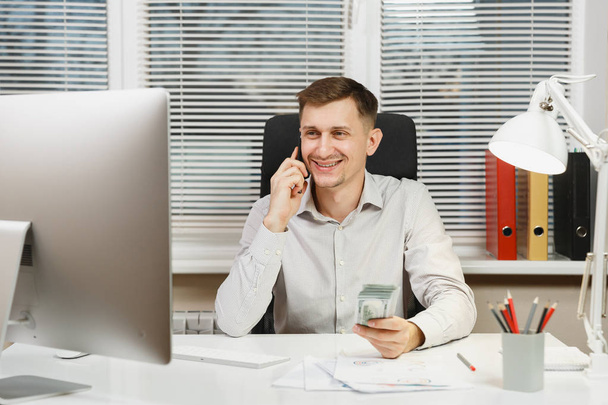 Handsome smiling business man in shirt sitting at the desk talking on mobile phone with lot of cash money, working at computer with modern monitor, lamp, documents in light office. Manager or worker. - Fotografie, Obrázek