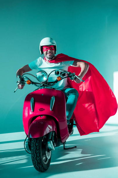smiling man in protective helmet, superhero mask and cape riding red scooter - 写真・画像