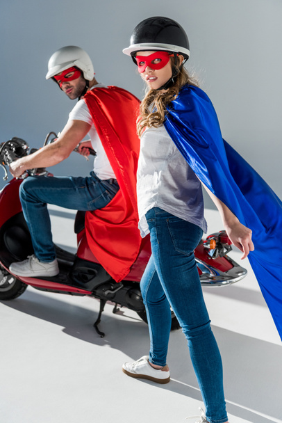 pareja con estilo en cascos y trajes de superhéroe con scooter rojo mirando a la cámara
 - Foto, Imagen