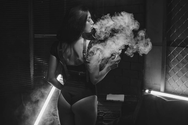 Young beautiful brunette woman smoking, vaping e-cigarette with smoke on black background. - 写真・画像