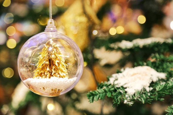 A little golden Christmas tree in the crystal clear bauble ball hangs as a decoration with blur background of Christmas tree with some snow on its leaf. - Photo, Image