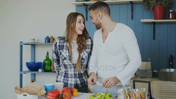 Laughing attractive woman surprising his boylfriend with a gift at home in the kitchen while he cooking breakfast - Metraje, vídeo