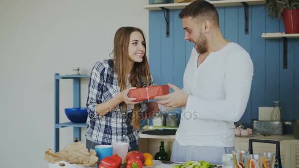 Cheerful woman surprising his boylfriend with birthday gift at home in the kitchen while he cooking breakfast - Filmagem, Vídeo