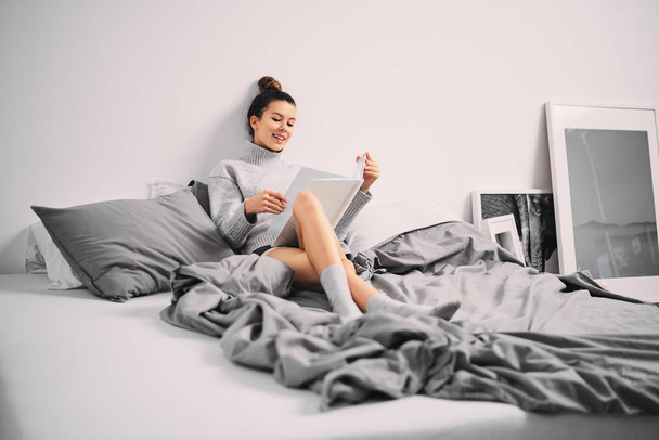 Chica sentada en la cama y leyendo libro en la mañana
 - Foto, Imagen