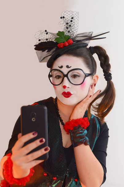 A young pretty girl mime, in a lady's hat and bright suit, makes selfie on her smartphone, grimacing, she parodies narcissism in women nowadays - Photo, Image