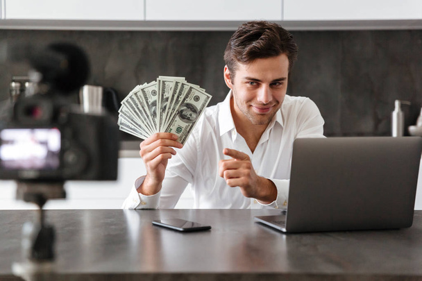 Smiling young man filming his video blog episode about new tech devices while sitting at the kitchen table with laptop and showing bunch of money banknotes - Фото, изображение