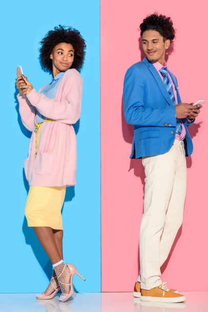 Young african amercian smiling couple with phones standing back to back on pink and blue background  - Photo, Image