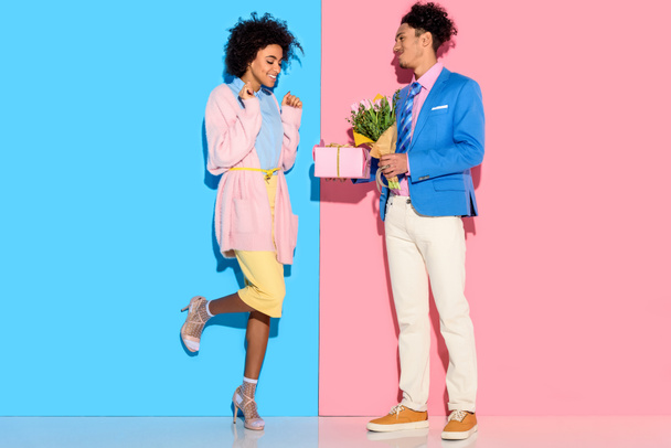 Hombre sonriente presentando caja y flores a la mujer sobre fondo rosa y azul
 - Foto, Imagen