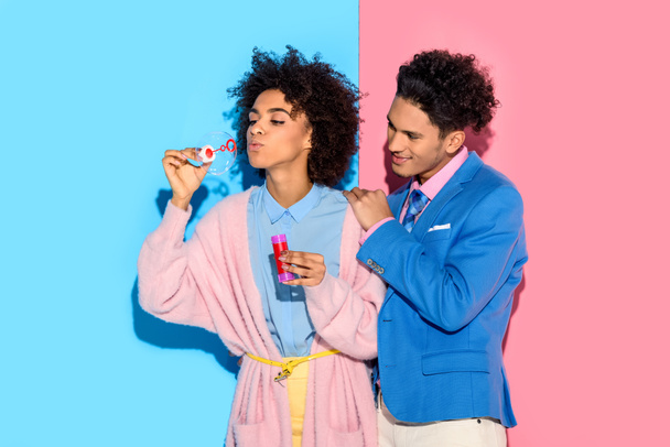 Afrikanisches Paar amüsiert sich mit Seifenblasen auf rosa und blauem Hintergrund - Foto, Bild