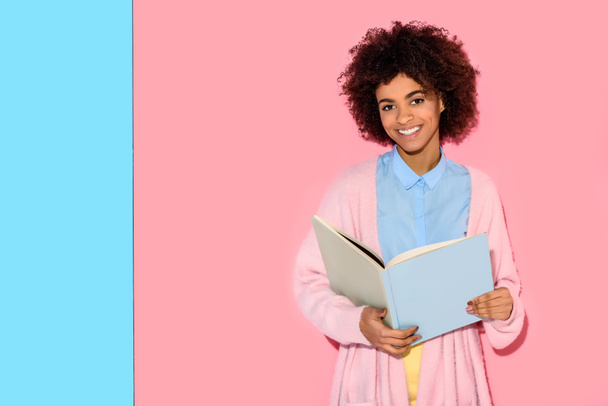 portrait de femme afro-américaine souriante avec un livre dans les mains regardant la caméra contre le mur rose et bleu
 - Photo, image