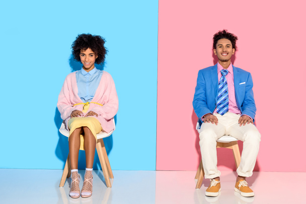 lachende Afrikaanse Amerikaanse echtpaar zitten op stoelen tegen roze en blauwe muur achtergrond - Foto, afbeelding