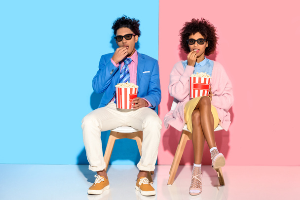 young african american couple sitting on chairs and eating popcorn against pink and blue wall - Photo, Image