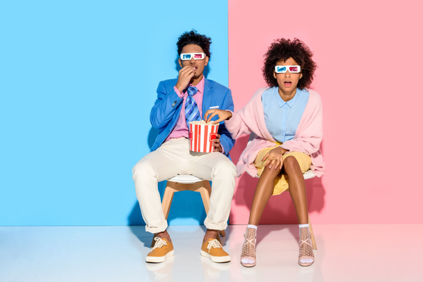 young african american couple sitting on chairs and eating popcorn against pink and blue wall - Fotoğraf, Görsel