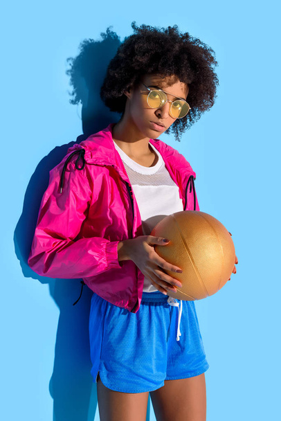  Young girl in glasses holding basketball ball and looking at camera on blue background     - Φωτογραφία, εικόνα