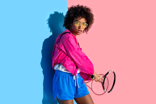 Africká americká dívka světlé houpačky tenisovou raketu na růžové a modré pozadí  - Fotografie, Obrázek