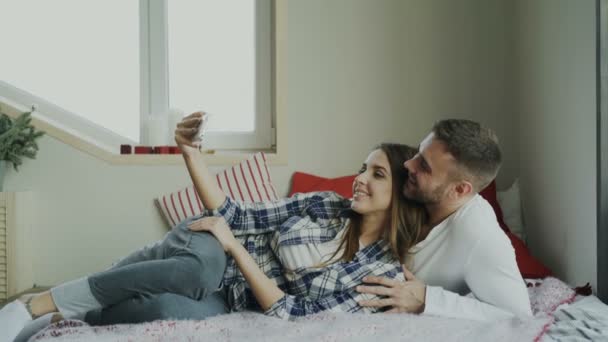 Cheerful cute and loving couple talking video call using smartphone camera and chatting with parents lying in bed at home - Video, Çekim