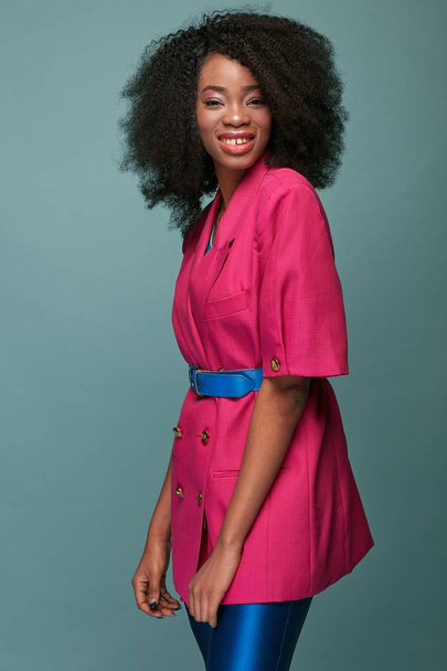 Beauty portrait of attractive young african girl with afro hairstyle. Smiling girl wearing pink jacket, blue leggings and belt posing on turquoise  background, . Studio shot. - Fotó, kép