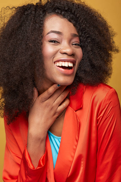 Kleurrijke close-up portret van jonge Afrikaanse meisje met afro kapsel. Lachende meisje dragen oranje jas en poseren op gele achtergrond. Studio schoot. - Foto, afbeelding