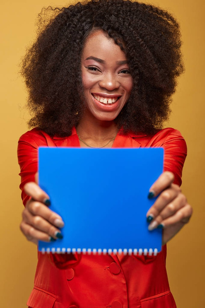 Colorful portrait of young african girl with afro hairstyle. Smiling girl wearing orange jacket holds blue copybook in her hands and posing on yellow background. Studio shot. - Fotografie, Obrázek