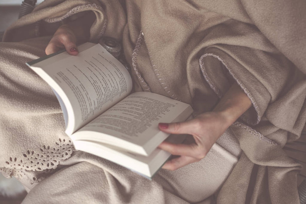 Νεαρή όμορφη γυναίκα κάθεται και διαβάζει ένα βιβλίο σε μια βεράντα που καλύπτεται με μια κουβέρτα και στο παρασκήνιο υπάρχει τζάκι - Φωτογραφία, εικόνα