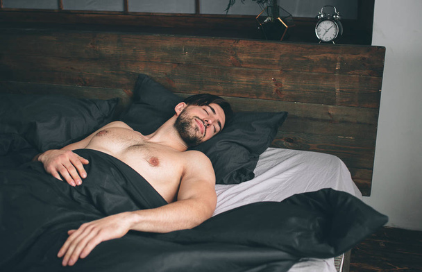Porträt eines fitten männlichen Modells, das im luxuriösen Schlafzimmer schläft, das in helles, warmes morgendliches Fensterlicht getaucht ist - Foto, Bild