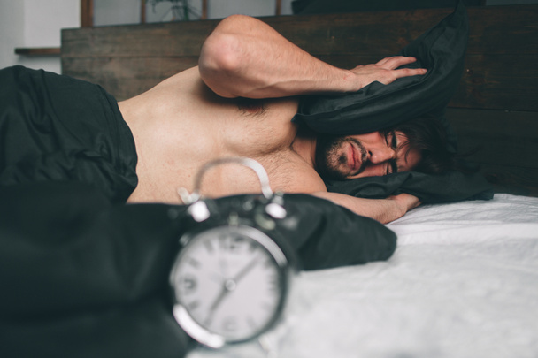 Пора просыпаться. Уставший человек в постели недоволен. Зрелый парень держит будильник во время проверки времени на работу
 - Фото, изображение