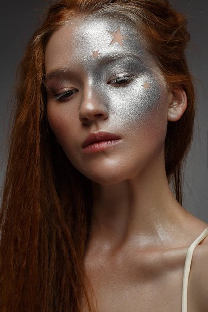 A young girl with red hair and a brilliant silver make-up with the stars. Beautiful model with shining skin. Holiday image. Beauty of the face. Photo taken in the studio. - Φωτογραφία, εικόνα
