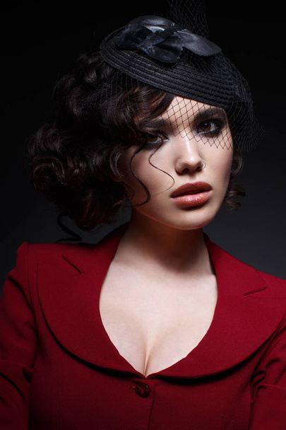 A young girl in a hat with a veil and with curls. A beautiful model in a red jacket with bright evening make-up. Shining skin. Beauty of the face. Photo taken in the studio. - Photo, Image