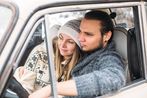 A Valentine couple sitting inside a snow retro car and kissing in a forest. Romantic Beauty girl in a hat and handsome man in a grey pullover. Horisontal picture - Photo, image