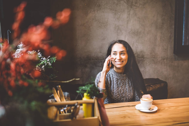 beautiful young brunette girl talking on mobile phone at wooden table near window and drinking coffee in cafe decorated with Christmas decor. Dressed in a gray knitted wool sweater - Foto, Imagem