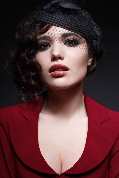 A young girl in a hat with a veil and with curls. A beautiful model in a red jacket with bright evening make-up. Shining skin. Beauty of the face. Photo taken in the studio. - Valokuva, kuva