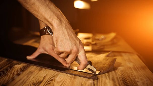 Working process of the leather belt in the leather workshop. Man holding tool. Tanner in old tannery. Wooden table background. Close up man arm. Warm Light for text and design. Web banner size - Zdjęcie, obraz