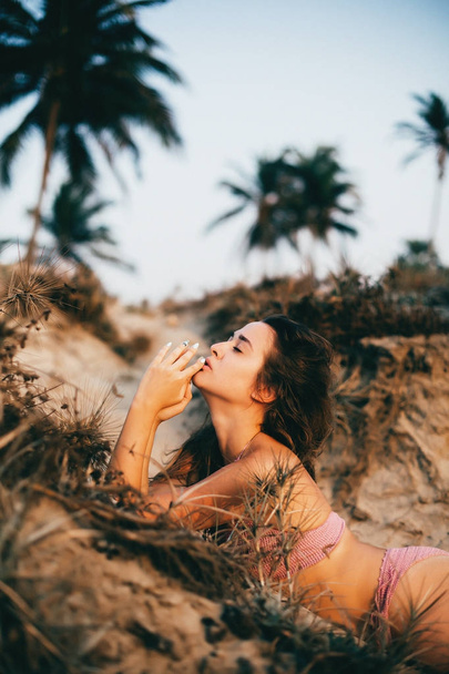 Όμορφη καυκάσιος γυναίκα με σέξι μαγιό στην παραλία στο ηλιοβασίλεμα φως του ήλιου, σέξι κορίτσι, γυναίκα στις διακοπές, ηλιοβασίλεμα στην παραλία, φοίνικες στην παραλία - Φωτογραφία, εικόνα