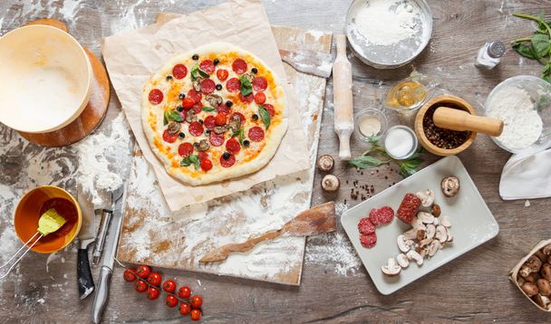 Top view cooking pizza with ingredients, tomatoes, salami and mushrooms on wooden tabletop - Foto, afbeelding