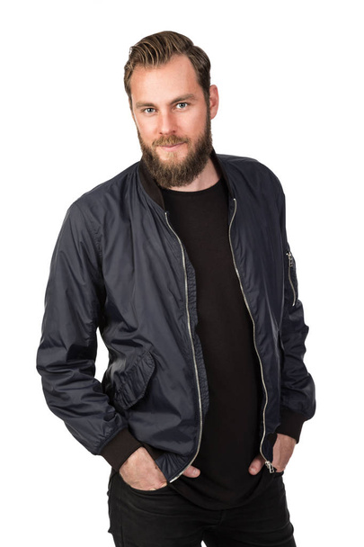 Handsome bearded man standing against a white background wearing a black tshirt and blue jacket in jeans. Looking at camera. - Фото, изображение