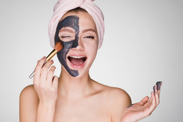 a funny young girl with white teeth, with a pink towel on her head, applies a clay mask to her face with a brush - Fotó, kép