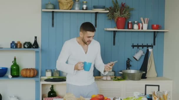 Slowmotion of Handsome young funny man dancing and singing in kitchen while surfing social media on his smartphone at home in the morning - Imágenes, Vídeo
