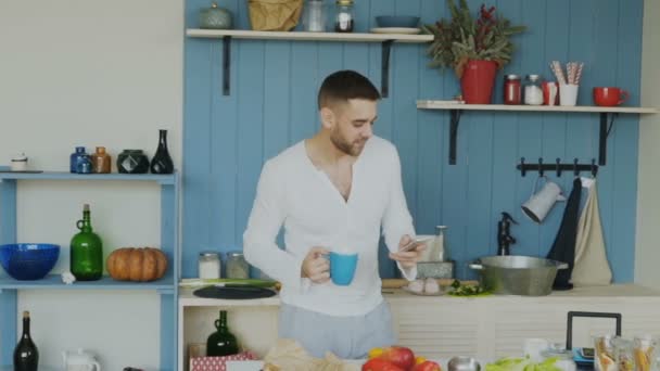 Zpomalený pohyb pohledný mladý legrační muž tančí a zpívá v kuchyni při surfování sociální média na jeho smartphone doma ráno - Záběry, video