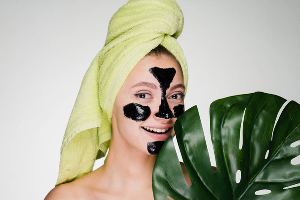 a smiling beautiful girl with a green towel on her head applied a black cleansing mask to the problem areas on her face - Photo, Image