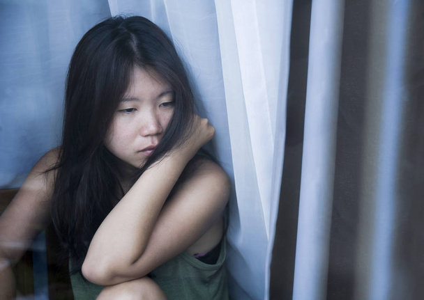 молодая грустная и подавленная азиатская китаянка смотрит вдумчиво через оконное стекло страдает болью и депрессией в концепции грусти
 - Фото, изображение
