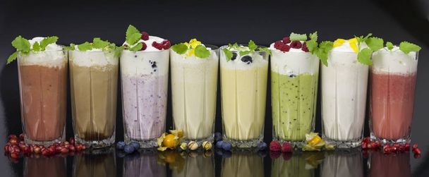 glasses of fresh milk cocktails (chocolate, strawberry, vanilla, kiwi, blueberry, pomegranate)  with fruits and berries on black background - Фото, изображение