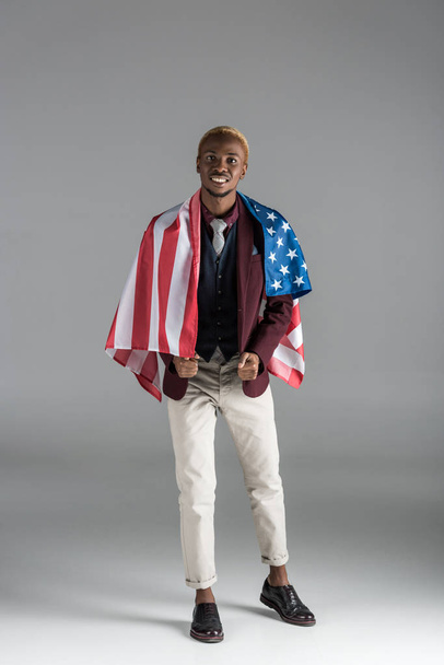 uśmiechnięty mężczyzna african american z amerykańską flagę na ramionach, patrząc na kamery na szarym tle  - Zdjęcie, obraz